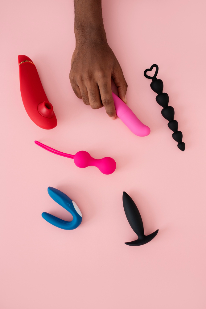 Mão de pessoa preta segurando diversos vibradores e briquedos eróticos em fundo rosa